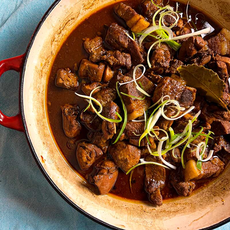 The recipe image of Filipeno Chicken and Pork Adobo made using Roni B's Adobo Sauce. The adobo is garnished with spring onions and it is a red cast iron casserole dish.