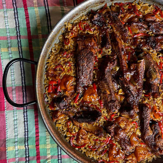 Recipe picture for Spanish Oven Baked Rice with Pork Ribs, Arroz al horno showing delicious roasted pork ribs nestled in flavourful rice in a round dish that's sat on a checkered table cloth.