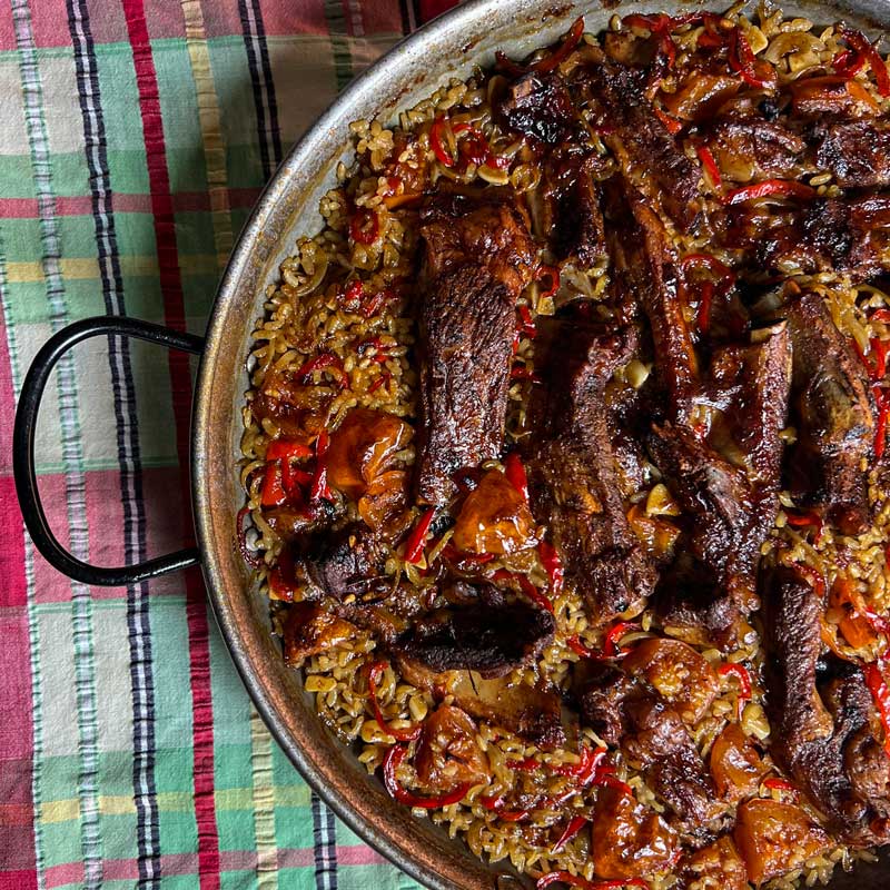 Recipe picture for Spanish Oven Baked Rice with Pork Ribs, Arroz al horno showing delicious roasted pork ribs nestled in flavourful rice in a round dish that's sat on a checkered table cloth.