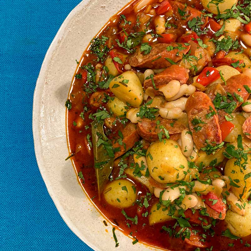 Image showing the recipe for Spanish Chorizo, white bean and potato stew garnished with chopped parsley. The stew is in a pale brown bowl on a blue table cloth.