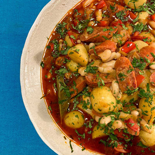 Image showing the recipe for Spanish Chorizo, white bean and potato stew garnished with chopped parsley. The stew is in a pale brown bowl on a blue table cloth.