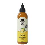 A 250ml squeeze bottle of All Dressed Up Miso Sesame dressing, featuring a black cap, amber-brown liquid inside, and a cream and yellow label with a sesame illustration.